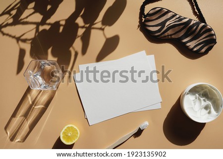 An empty sheet of white paper surrounded by container with cream, a lemon, tooth brush, sleeping mask and a glass on yellow surface, a mockup            