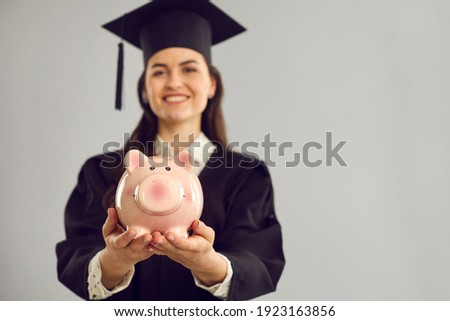 Close up of a pink piggy bank in the hands of a joyful female student standing on a gray background. Concept of a budget form of education, college or university payments and scholarships. Blur. Royalty-Free Stock Photo #1923163856