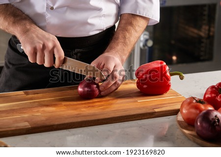 Cropped male chef precisely slicing red onions on wooden cutting board in restaurant kitchen, close-up photo of male hands holding knife. food, dish, cooking concept