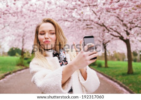 Cute young woman gesturing peace sign while taking her picture with mobile phone. Caucasian female model at spring blossom park taking self portrait with smart phone.