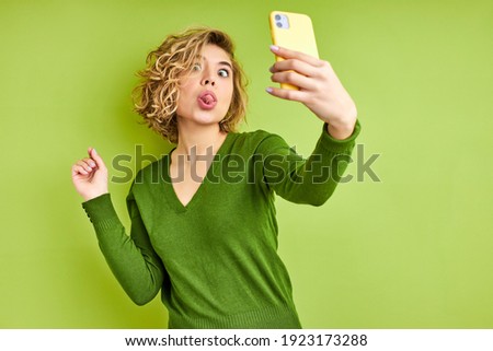 woman going crazy, taking selfie photo on mobile phone, wearing everyday stylish green outfit isolated over colorful wall background looking at device screen