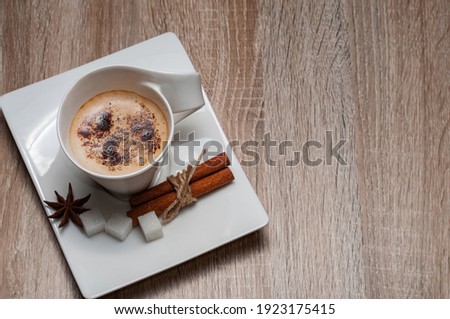 White cup with coffee.The cup is on a white saucer, next to it are three lumps of sugar, cinnamon sticks and and star anise.Whipped coffee and sprinkled with grated chocolate.Copy space.