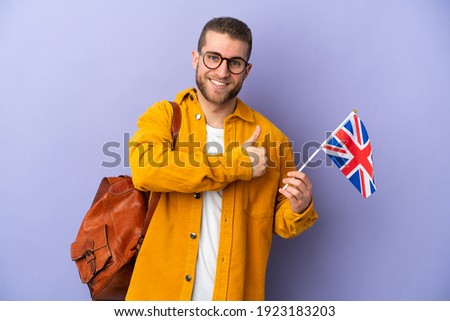 Young caucasian man holding an United Kingdom flag isolated on purple background giving a thumbs up gesture
