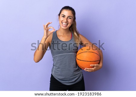 Young hispanic woman over isolated purple background playing basketball and making OK sign