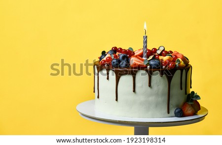 Birthday party sweet cake with chocolate drips and fresh fruits Raspberry strawberry blueberry cranberry. Cake with lighted candle on light yellow background
