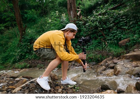 Young male cameraman with a camera on a stabilizer in his hands shoots a video of nature on a hike. Guy stands with a camera on a gimbal near a mountain stream and makes content.