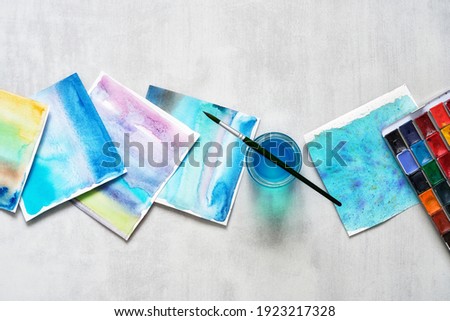 Set of paper sheets of watercolor patterns, glass of water, brush for painting and watercolor paints. Top view.