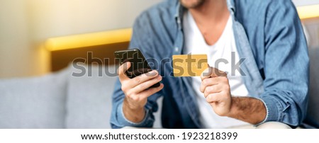 Panoramic photo of credit card and smartphone in male hands. Caucasian guy, uses smartphone and credit card for online payment of shopping or delivery of goods, sit at home on sofa, enter card details