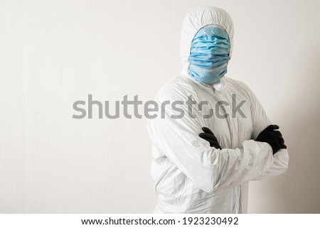 a man in a protective suit hung with medical masks posing against a wall background showing various gestures with his fingers