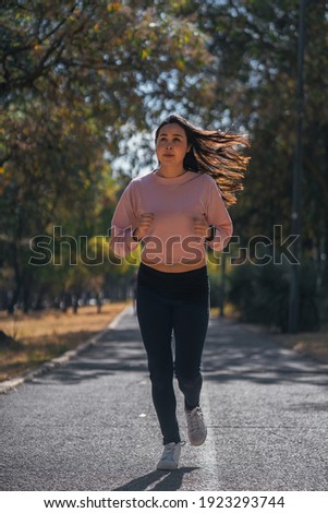 pretty Hispanic woman running at sunrise on urban park track. warm colors and green nature background- wears black pants, pink sweater and white tennis shoes