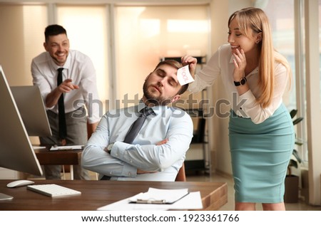 Young woman sticking note with word Fool to colleague's face in office. Funny joke Royalty-Free Stock Photo #1923361766