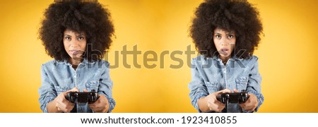 Young mixed african american woman, playing video games with surprised face, with game controller, fun, on orange yellow background
