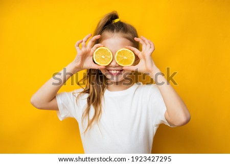 Little smiling cute blond girl in yellow t-shirt holding halves of fresh sour lemon fruit near eyes and showing tongue over yellow background. Healthy lifestyle and clean eating concept Royalty-Free Stock Photo #1923427295