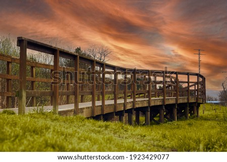 Brown wooden walkway that passes over a stream in nature surrounded by vegetation like grass. and a dirt road. sunset orange