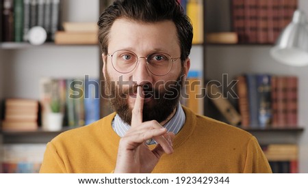 Shh, man secret finger. Suspicious bearded man in glasses in office or apartment room looking at camera and brings his index finger to his mouth lips and she say shhh. Close-up view