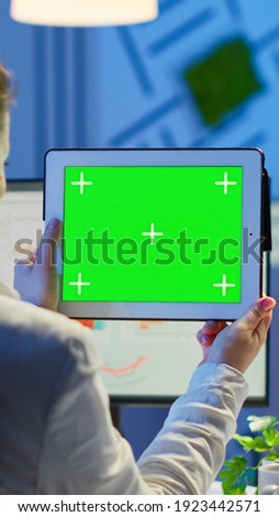 Businesswoman holding tablet with green screen monitor sitting at desk in business office late at night. Freelancer watching desktop monitor display with green mockup, chroma key working overtime.