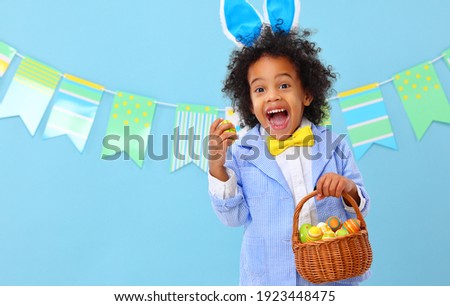 Amazed cute little African American boy in stylish suit and bunny ears holding Easter basket with colorful eggs and screaming happily against blue background with party flags