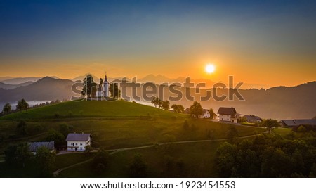 Skofja Loka, Slovenia - Aerial panoramic view of amazing golden sunrise at the beautiful hilltop church of Sveti Tomaz (Saint Thomas) with and the Julian Alps at background at summer time