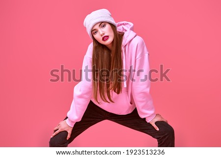 Youth fashion. Modern teen girl in a pink sweatshirt and a hat posing at studio on a pink background. Cosmetics and make-up.
