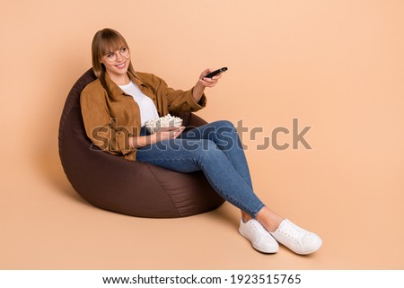 Full size photo of attractive happy pretty young lady switch channel sit bean bag hold pop corn isolated on beige color background