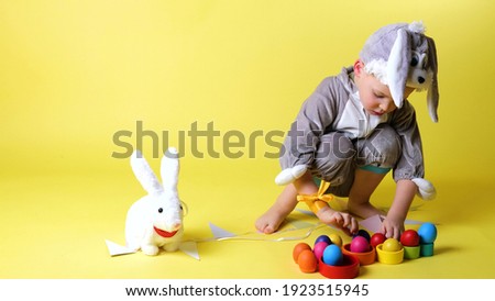 little cheerful boy in bunny grey costume with bunny ears playing with toy rabbit, colorful eggs on trendy ultimate yellow background, slow motion, Happy And Fun Easter Day concept celebrate. 