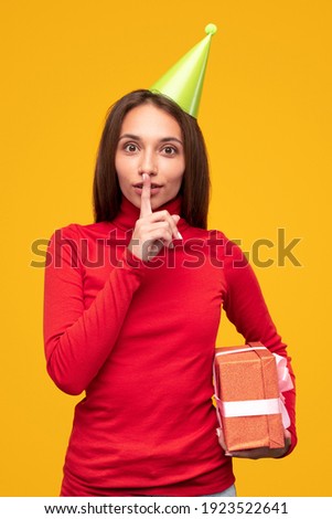 Young female with gift box making silence gesture and asking to keep secret during birthday celebration against yellow background