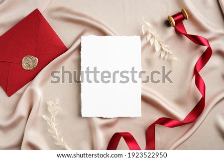 Elegant wedding stationery set top view. Flat lay blank invitation card mockup, red ribbon, envelope on beige satin.