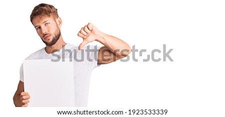 Young caucasian man holding blank empty banner with angry face, negative sign showing dislike with thumbs down, rejection concept 