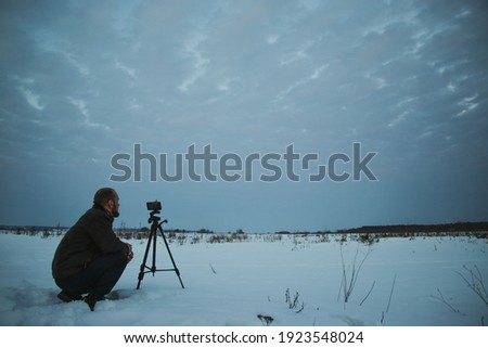 Man with phone on tripod making time-lapse photographs of winter zidio sunset. The field, road and trees are covered with snow.