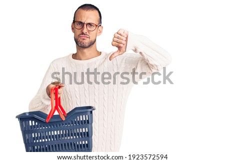 Young handsome man holding supermarket shopping basket with angry face, negative sign showing dislike with thumbs down, rejection concept 