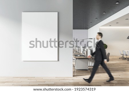 Businessman goes to blank white poster on light grey wall in modern open space office with light furniture and wooden floor. Mock up Royalty-Free Stock Photo #1923574319
