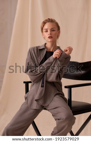 studio portrait of young elegant woman in grey linen costume and black high heels sitting at black high chair against beige textile background.