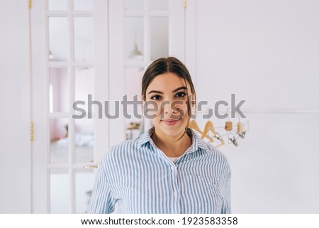 Authentic portrait of attractive caucasian woman in shirt, smiling and looking on camera in white modern room.
