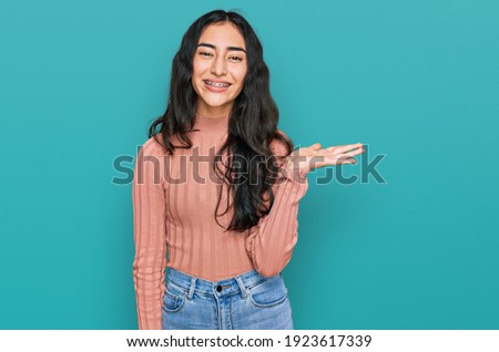 Hispanic teenager girl with dental braces wearing casual clothes smiling cheerful presenting and pointing with palm of hand looking at the camera. 