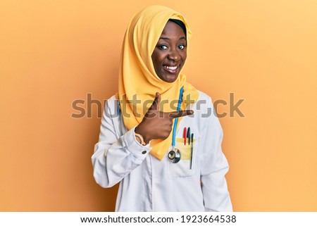 Beautiful african young woman wearing doctor uniform and hijab cheerful with a smile on face pointing with hand and finger up to the side with happy and natural expression 