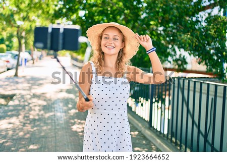 Young beautiful caucasian woman with blond hair smiling happy outdoors taking a selfie picture using selfie stick