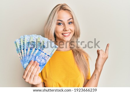 Beautiful caucasian blonde girl holding south african 100 rand banknotes smiling happy pointing with hand and finger to the side 