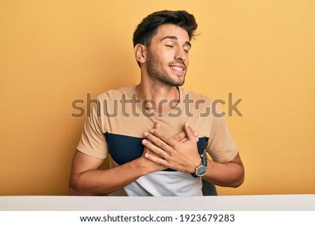 Young handsome man wearing casual clothes sitting on the table smiling with hands on chest, eyes closed with grateful gesture on face. health concept. 