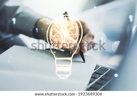 Creative light bulb with human brain hologram and hand writing in notepad on background with laptop, artificial Intelligence and neural networks concept. Multiexposure