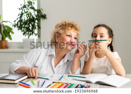 Happy grandchild with grandmother having fun, drawing colored pencils, sitting together at home, laughing preschool girl with smiling grandma painting picture, playing with granddaughter