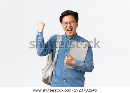 University, study abroad and lifestyle concept. Happy rejoicing asian male student with braces triumphing, pass exams, finish final semester, fist pump and shouting yes with satisfaction, hold laptop Royalty-Free Stock Photo #1923702245
