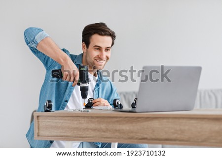 Assemble furniture with own hands and modern home renovation during covid. Happy handsome young male screwing nuts with screwdriver and watch video instruction in laptop, at home workroom, free space Royalty-Free Stock Photo #1923710132
