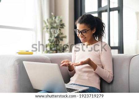 Positive smart young African American female freelancer or a student in glasses sitting on the couch with laptop on the lap, studying from online webinar or a course at home, video calling and talking Royalty-Free Stock Photo #1923719279
