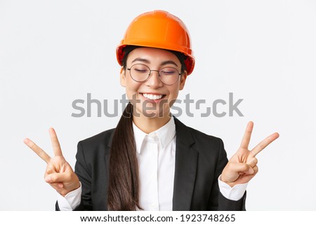 Close-up of cheerful successful female asian engineer, construction architect in safety helmet and business suit showing peace signs and smiling kawaii, standing white background