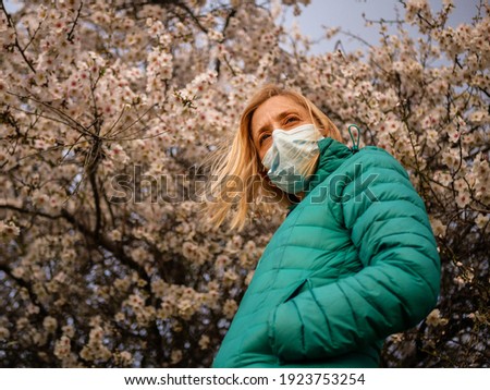 Blond woman with blue eyes, wearing a green winter coat and a mask, posing under a flowering almond tree