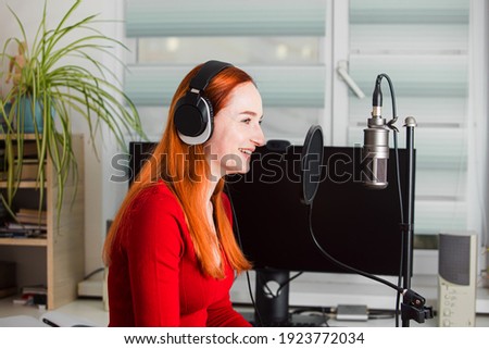 The young woman in headphones is sitting in front of a professional microphone and talking. The woman with red hair in a red dress on the background of the computer
