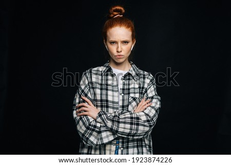 Disgruntled red-haired young woman wearing wireless earphones with crossed hands looking at camera on isolated black background. Pretty redhead lady model emotionally showing facial expressions.