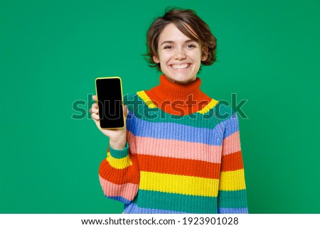 Smiling cheerful young brunette woman 20s years old wearing casual colorful sweater standing hold mobile cell phone with blank empty screen isolated on bright green color background studio portrait