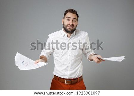 Confused puzzled young bearded business man 20s wearing classic white shirt hold papers document spreading hands isolated on grey color background studio portrait. Achievement career wealth concept