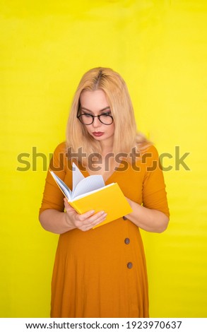 Beautiful blonde woman in glasses holding book in her hands over yellow background